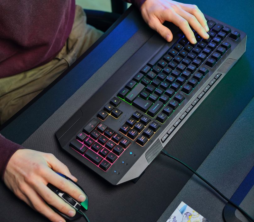 Manos de una persona utilizando un Teclado Gaming Iluminado Trust GXT 836 Evocx y un mouse, sobre una mesa con superficie oscura.