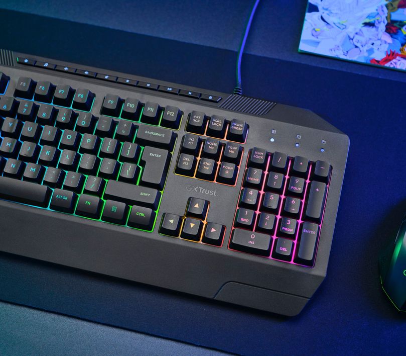 Teclado Gaming Iluminado Trust GXT 836 Evocx sobre una mesa con luz azulada, junto a un mouse con luces y papeles desordenados al fondo.