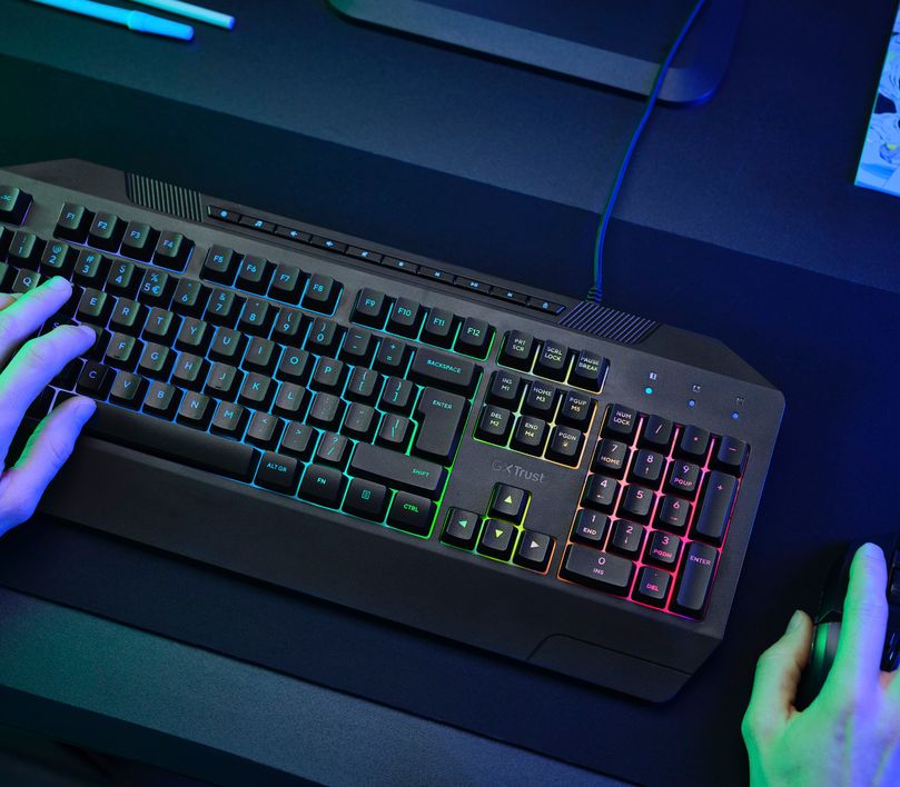 La imagen muestra un Teclado Gaming Iluminado Trust GXT 836 Evocx sobre un escritorio con iluminación azul. También se ve una mano sobre el mouse y otra en las teclas WASD.
