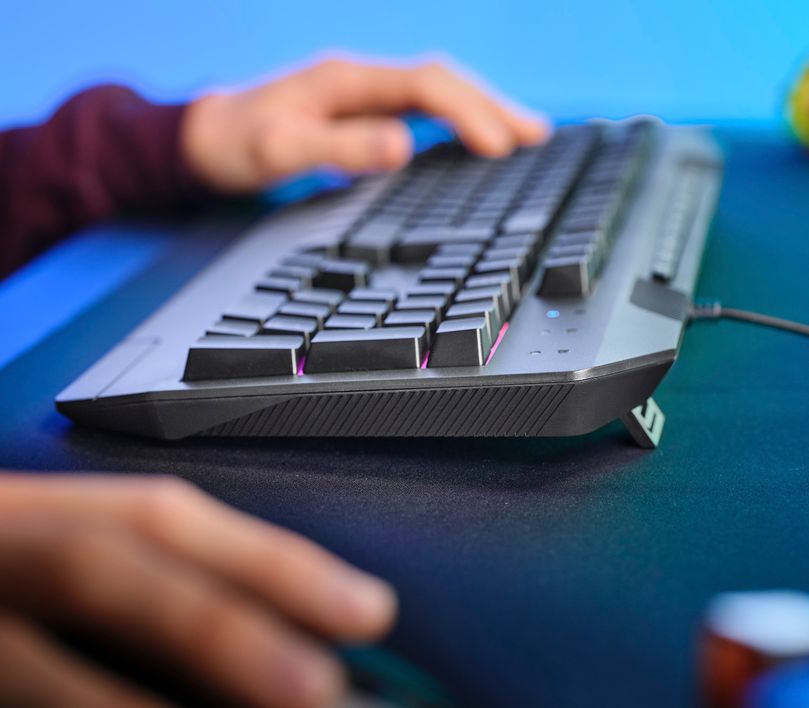 La imagen muestra un Teclado Gaming Iluminado Trust GXT 836 Evocx. Las manos de una persona están tecleando sobre el teclado, que se encuentra sobre un escritorio con fondo azul.