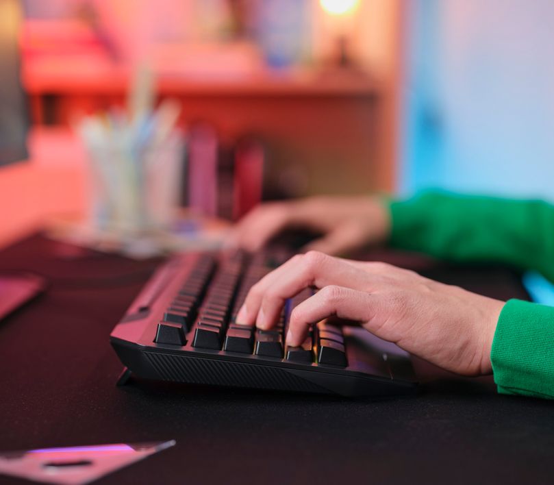 Una persona está utilizando un Teclado Gaming Iluminado Trust GXT 836 Evocx en un escritorio con iluminación tenue y colores cálidos en el fondo.