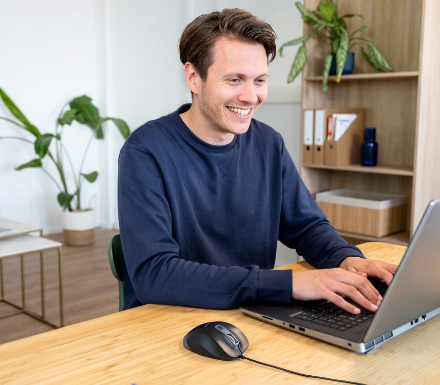 Una persona sentada frente a una computadora portátil en un escritorio de madera, utilizando un Mouse ergonómico Trust Fyda con una planta y estantería al fondo.
