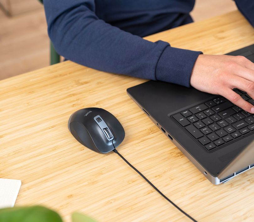 Una persona utilizando una computadora portátil con un Mouse ergonómico Trust Fyda conectado, sobre una mesa de madera.