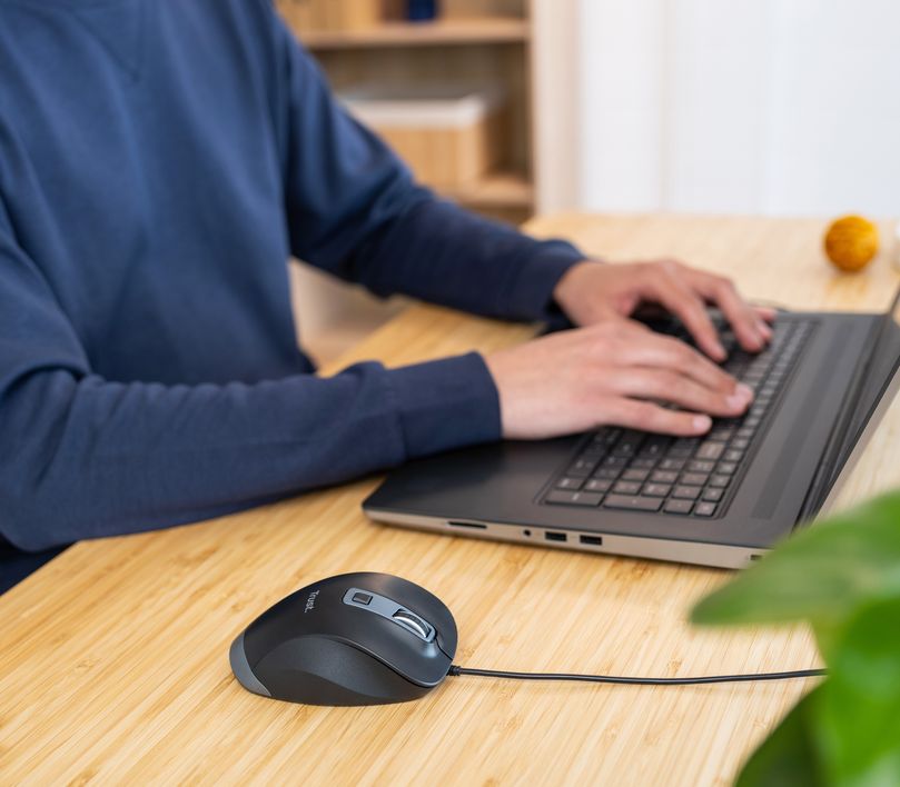 Una persona está utilizando una computadora portátil sobre un escritorio de madera, con un Mouse ergonómico Trust Fyda al lado.