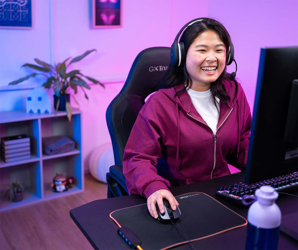 La imagen muestra a una persona sentada frente a una computadora. Tiene una mano en el teclado y la otra en un Mouse gaming Trust GXT 922 Ybar de color negro. La iluminación alrededor es de tonos azules y violetas.