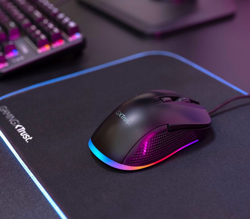 Mouse gaming Trust GXT 922 Ybar de color negro sobre un mousePad con iluminación LED multicolor, junto a un teclado parcialmente visible en el fondo, todo sobre un escritorio con una superficie oscura.
