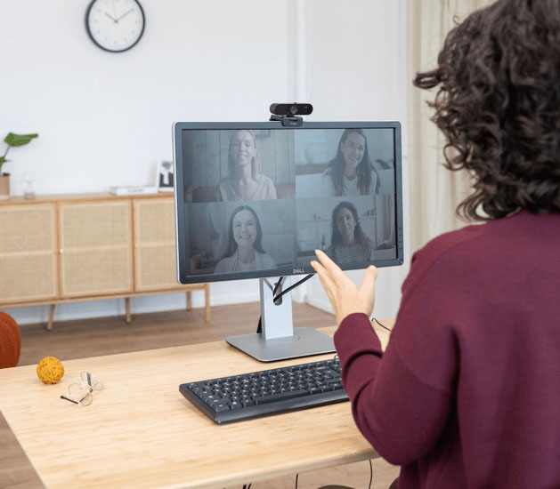 Persona participando en una videollamada grupal desde una computadora de escritorio, con una Taxon QHD Webcam de Trust acoplada al monitor, en un entorno hogareño bien iluminado y ordenado.