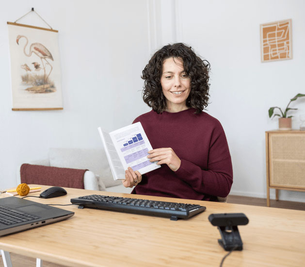 Mujer mostrando un libro frente a una computadora portátil y una Taxon QHD Webcam de Trust, en una videollamada desde un entorno hogareño luminoso y profesional.