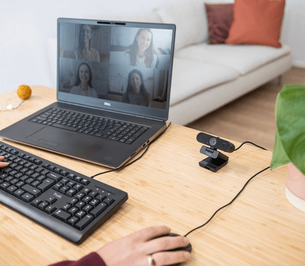 Videollamada grupal en una notebook conectada a una Taxon QHD Webcam de Trust colocada sobre el escritorio, junto a un teclado Trust y mouse, en un entorno hogareño ordenado.