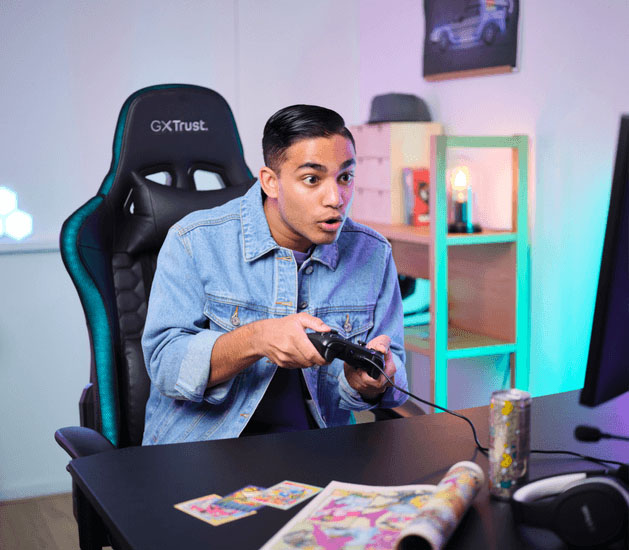 Joven concentrado juega en silla Trust GXT negra y azul, sosteniendo el controlador Muta GXT 541 de Trust, ideal para vós que jugás en PC.