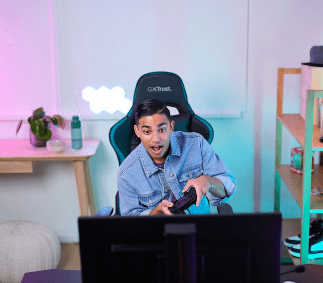 Gamer emocionado usando el controlador Muta GXT 541 de Trust, sentado en silla gaming, con setup de luces neón.