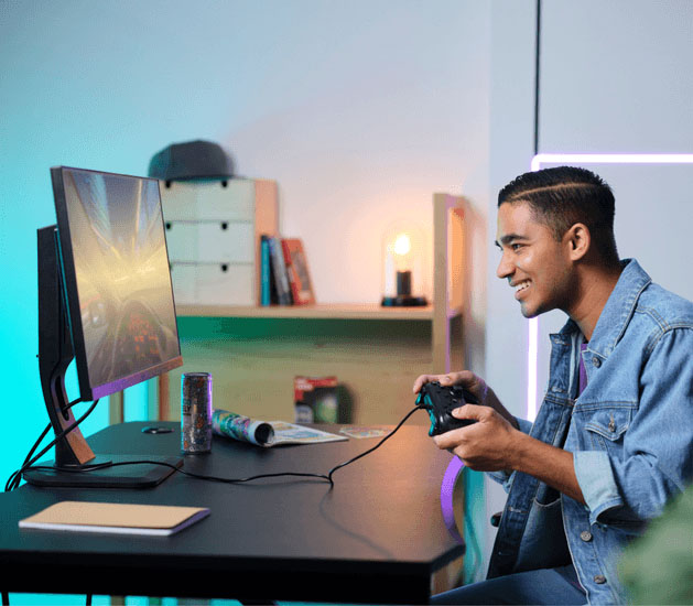 Joven sonriente juega con el controlador Trust Muta GXT 541 negro en su PC, concentrado en la pantalla y con una bebida energizante en el escritorio.