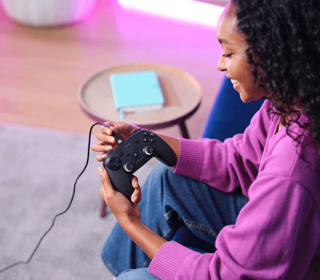 Joven sonriente sujeta un controlador Trust GXT 542 Muta negro con cable, jugando en un entorno interior con luz rosa.