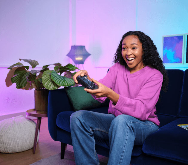 Mujer joven, emocionada, sentada en un sofá azul, usando el controlador de juego Trust GXT 542 Muta en un living iluminado con tonos púrpura y azul neón.