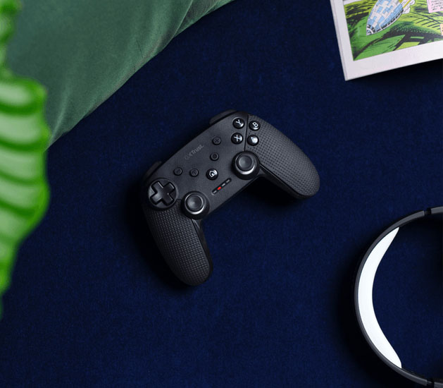 Primer plano del controlador Trust GXT 542 Muta negro, posado sobre una superficie azul oscura, mostrando los botones, joysticks y la textura de agarre en un plano cenital.