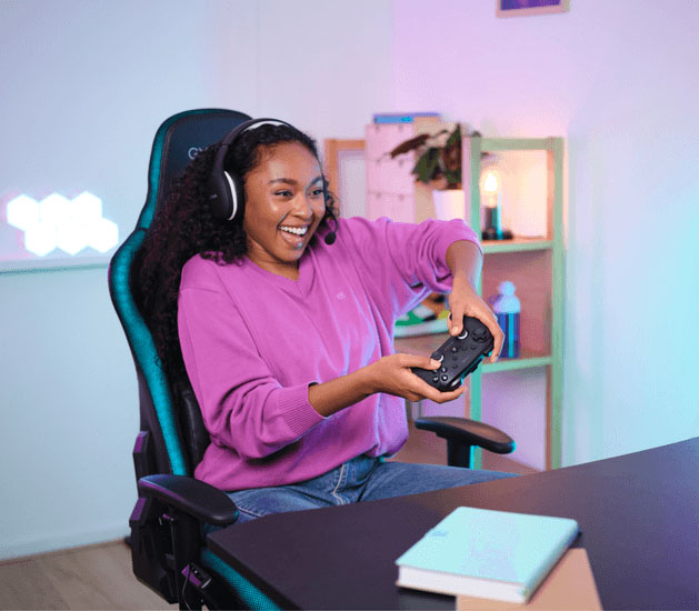 Chica usando audífonos y el controlador Trust GXT 542 Muta, sentada en una silla gamer negra en una habitación iluminada con luces de neón rosa y azul.