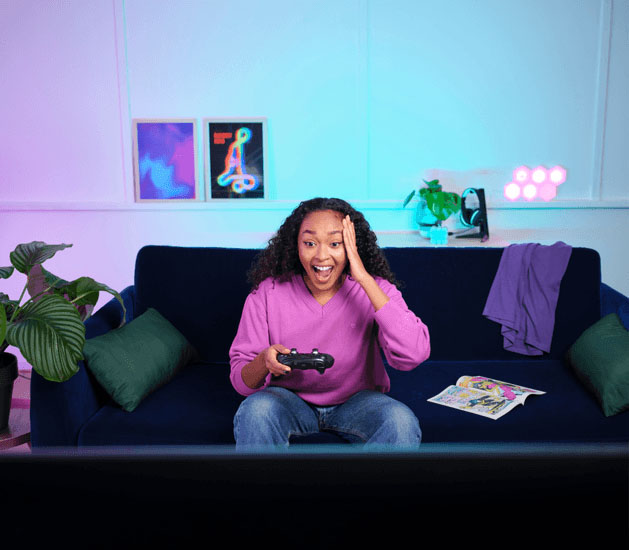 Gamer emocionada con una expresión de sorpresa, sentada en un sofá azul, sujetando el control Trust GXT 542 Muta mientras mira la pantalla.