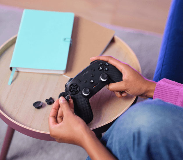 Manos intercambiando los D-pads del controlador Trust GXT 542 Muta negro, mostrando los accesorios y el control sobre una mesa de madera.