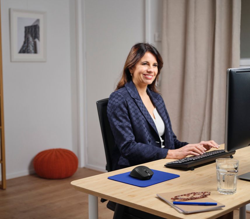 Una persona sentada en un escritorio de oficina, trabajando en una computadora. En el escritorio hay un vaso de agua, un cuaderno con un bolígrafo azul, y un Mouse inalámbrico multidispositivo Trust Ozaa+ de color Negro sobre una alfombrilla azul. Al fondo, se puede ver una calabaza decorativa y una fotografía enmarcada en la pared.