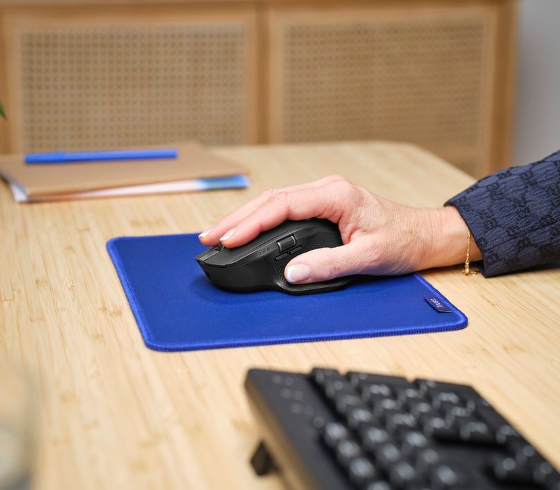 Una mano usando un Mouse inalámbrico multidispositivo Trust Ozaa+ de color Negro sobre una alfombrilla azul en un escritorio de madera clara. Se puede ver también un teclado negro y unos cuadernos con un bolígrafo azul encima.
