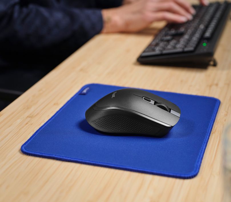 Un Mouse inalámbrico multidispositivo Trust Ozaa+ de color Negro sobre una alfombrilla azul en un escritorio de madera clara. Al fondo, una persona está escribiendo en un teclado.