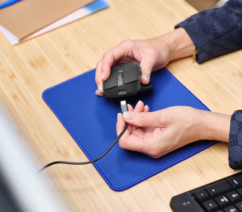 Una persona está conectando un cable USB-C a un Mouse inalámbrico multidispositivo Trust Ozaa+ de color Negro sobre una alfombrilla azul. La escena se desarrolla en un escritorio de madera clara, donde también se observa un teclado y algunos documentos.