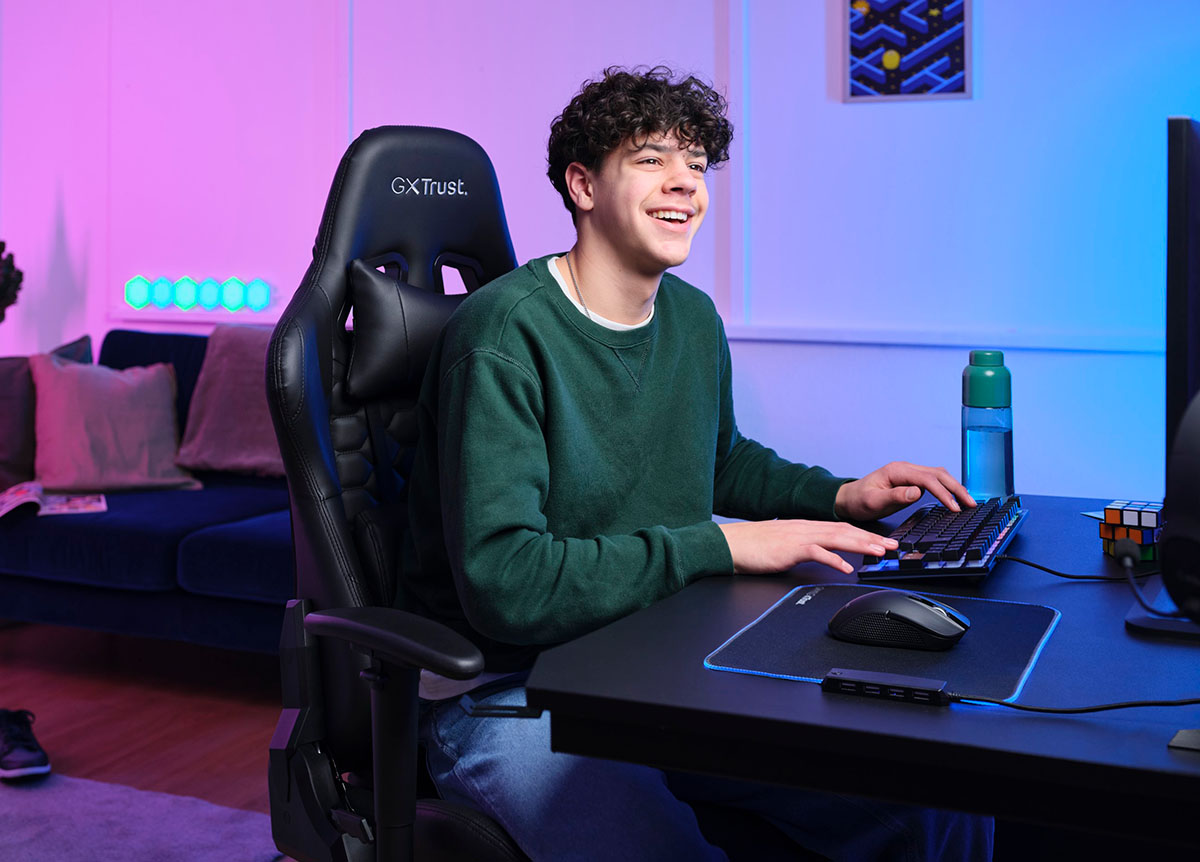 La imagen muestra a una persona sentada frente a una computadora. Tiene una mano en el teclado y la otra en un Mouse gaming inalámbrico Trust GXT 923 Ybar. La iluminación alrededor es de tonos azules y violetas.