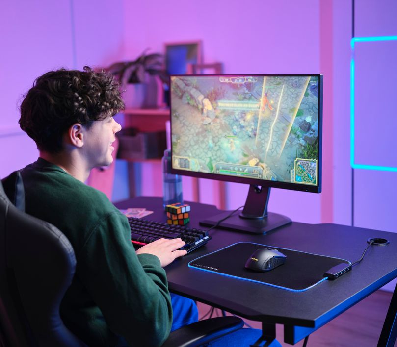 Una persona está jugando a un videojuego en una computadora con una pantalla grande. La habitación está iluminada con luces de colores rosa y azul. El teclado y el Mouse gaming inalámbrico Trust GXT 923 Ybar también son visibles en la imagen.