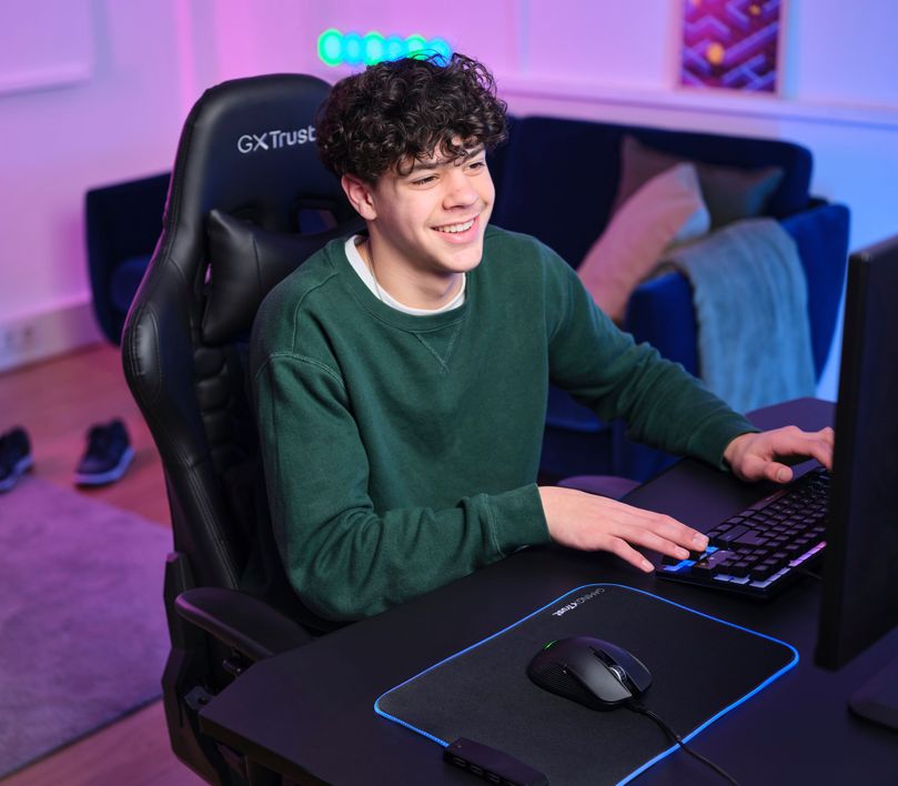La imagen muestra a una persona con un suéter verde sentada frente a una computadora. Está utilizando un teclado y un Mouse gaming inalámbrico Trust GXT 923 Ybar sobre un mousepad con iluminación LED. El ambiente tiene una iluminación azul y violeta, creando un espacio de juego moderno y tecnológico.