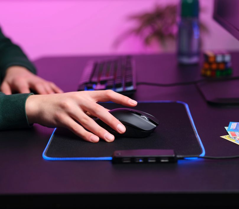Una mano descansa sobre un Mouse gaming inalámbrico Trust GXT 923 Ybar. El mouse está situado en un mousepad con iluminación LED azul, sobre un escritorio junto a un teclado mecánico. En el fondo, se ven elementos desenfocados como una botella y un cubo de Rubik.