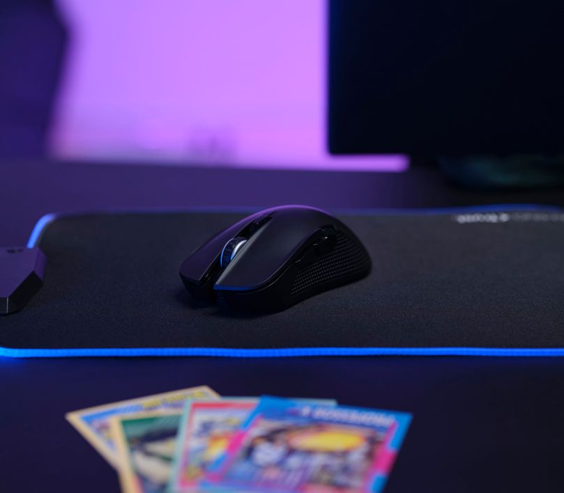 Mouse gaming inalámbrico Trust GXT 923 Ybar sobre un mousepad con iluminación LED azul, junto a tarjetas coleccionables desenfocadas y un monitor de computadora al fondo en un ambiente oscuro con luces violetas.