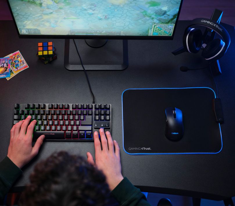 La imagen muestra una vista aerea de una persona en un escritorio de gaming con un teclado mecánico RGB, un Mouse gaming inalámbrico Trust GXT 923 Ybar sobre un mousepad con iluminación LED azul, un monitor encendido mostrando un juego, auriculares colgados y un cubo de Rubik al lado.