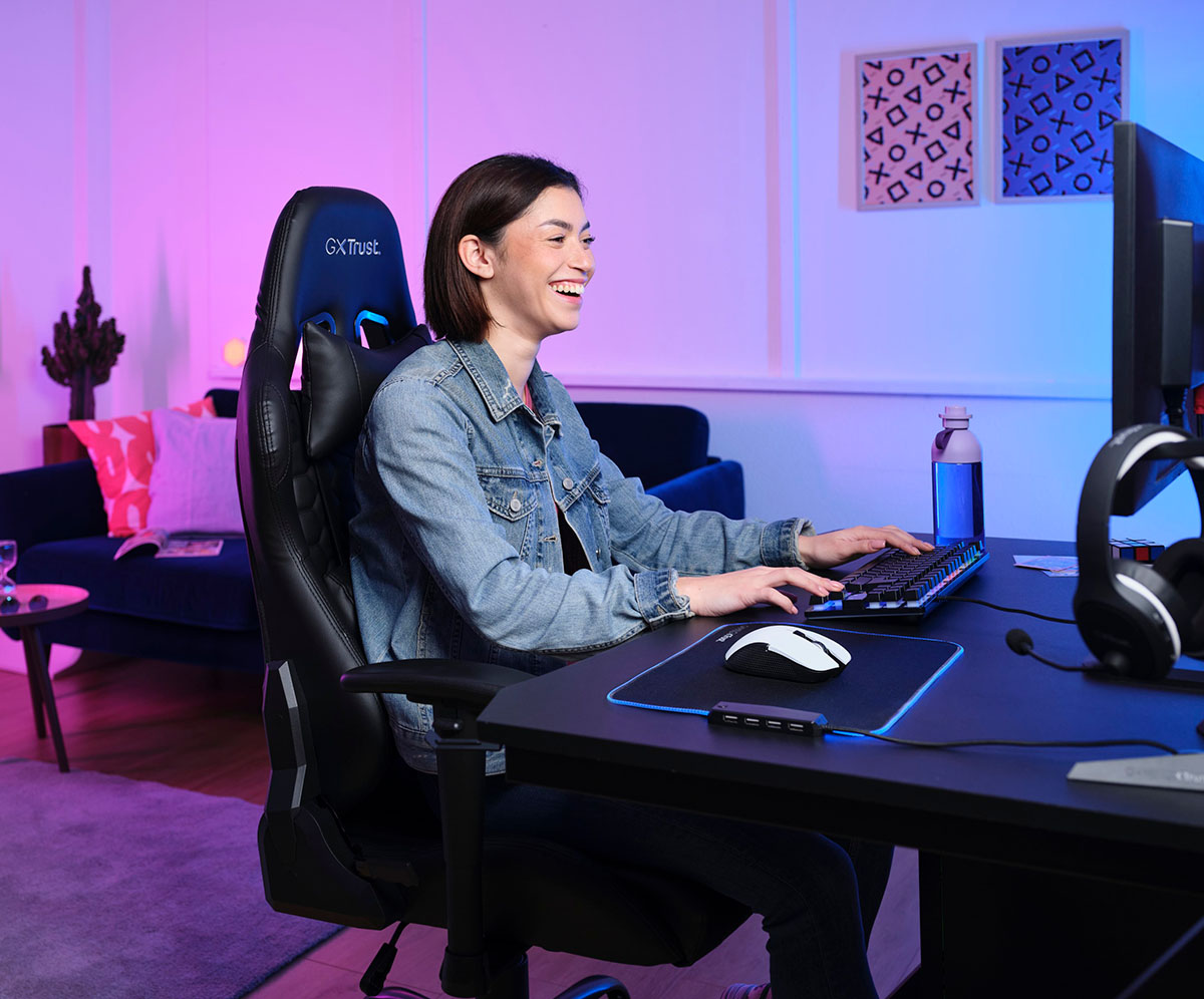 La imagen muestra a una persona sentada frente a una computadora. Tiene una mano en el teclado y hay un Mouse gaming inalámbrico Trust GXT 923W Ybar color blanco sobre un mousepad. La iluminación alrededor es de tonos azules y violetas.