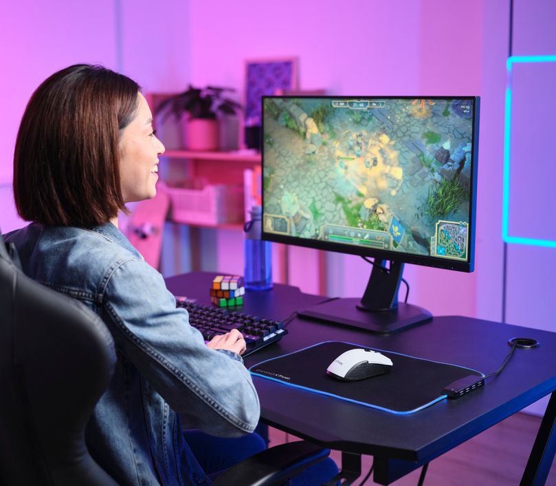 Una persona está jugando a un videojuego en una computadora con una pantalla grande. La habitación está iluminada con luces de colores rosa y azul. El teclado y el Mouse gaming inalámbrico Trust GXT 923W Ybar color blanco también son visibles en la imagen.