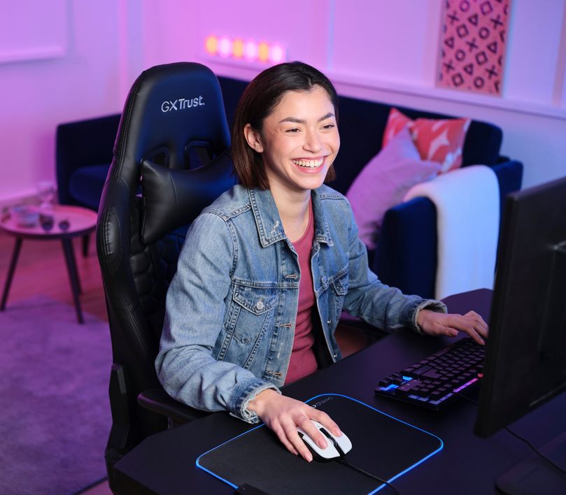 La imagen muestra a una persona sentada frente a una computadora. Está utilizando un teclado y un Mouse gaming inalámbrico Trust GXT 923W Ybar color blanco sobre un mousepad con iluminación LED. El ambiente tiene una iluminación azul y violeta, creando un espacio de juego moderno y tecnológico.
