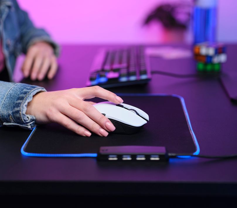Una mano descansa sobre un Mouse gaming inalámbrico Trust GXT 923W Ybar color blanco. El mouse está situado en un mousepad con iluminación LED azul, sobre un escritorio junto a un teclado mecánico. En el fondo, se ven elementos desenfocados como una botella y un cubo de Rubik.