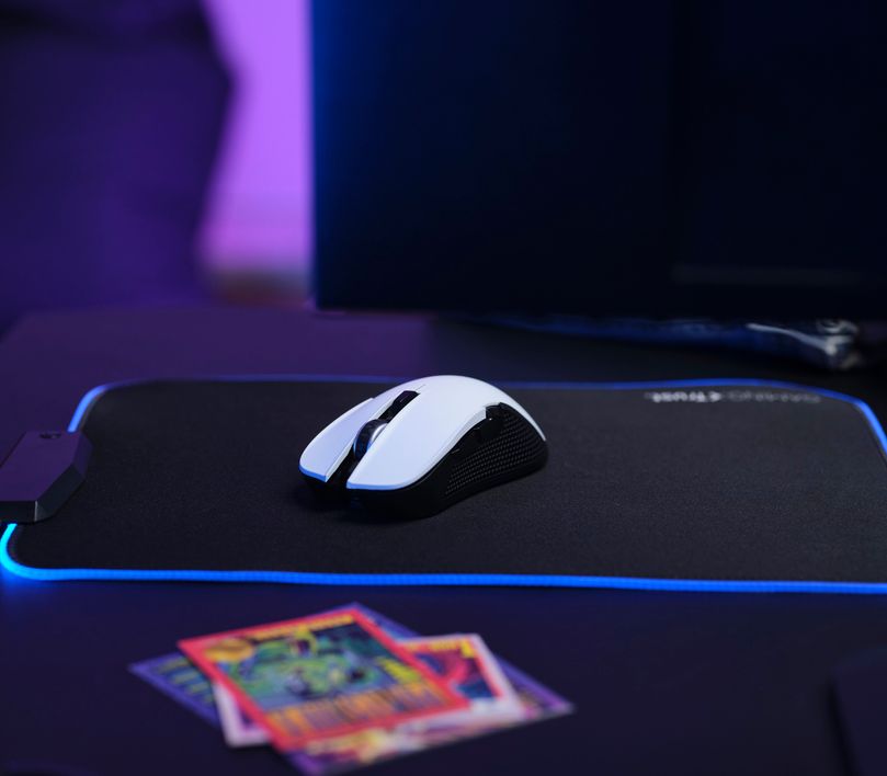 Mouse gaming inalámbrico Trust GXT 923W Ybar color blanco sobre un mousepad con iluminación LED azul, junto a tarjetas coleccionables desenfocadas y un monitor de computadora al fondo en un ambiente oscuro con luces violetas.