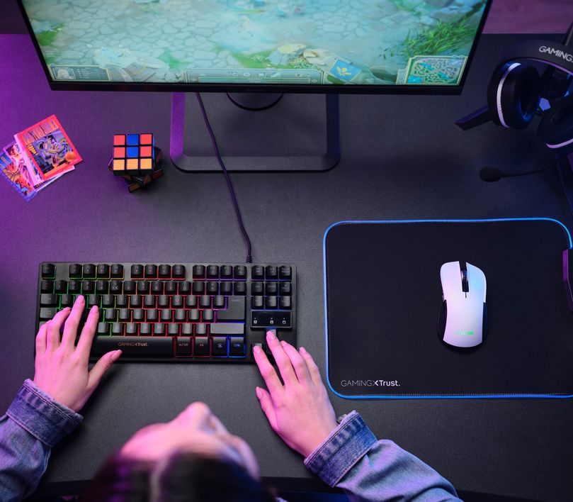 La imagen muestra una vista aerea de una persona en un escritorio de gaming con un teclado mecánico RGB, un Mouse gaming inalámbrico Trust GXT 923W Ybar color blanco sobre un mousepad con iluminación LED azul, un monitor encendido mostrando un juego, auriculares colgados y un cubo de Rubik al lado.
