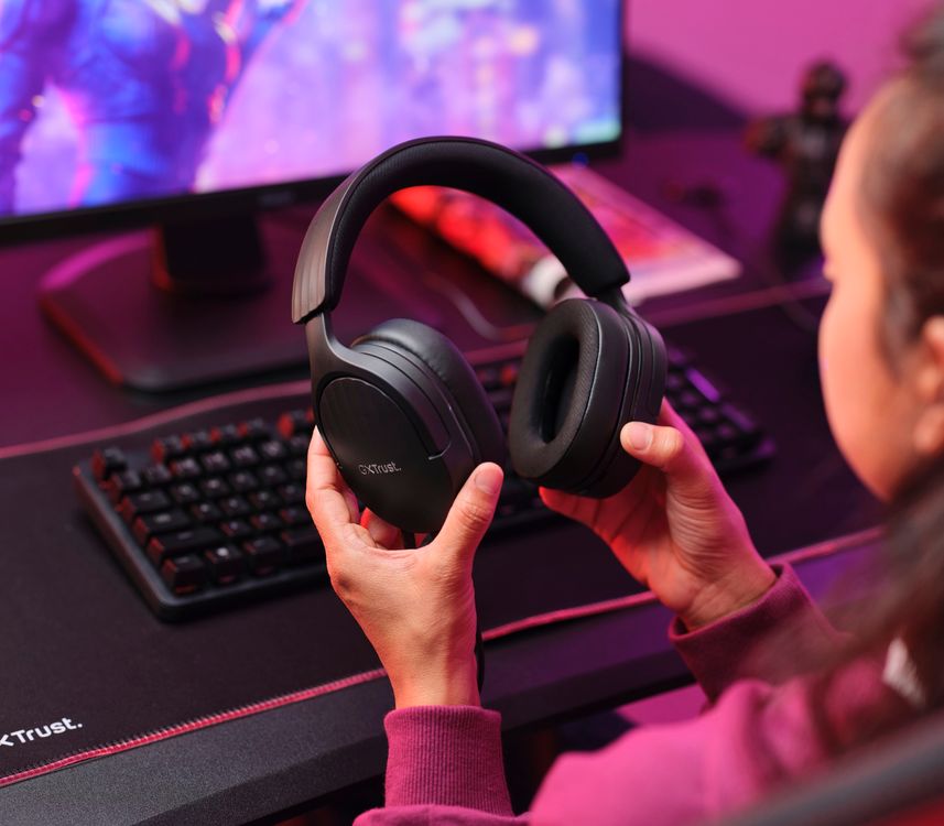 La imagen muestra una persona sosteniendo en sus manos unos Auriculares Gaming Multiplataforma Trust GXT 489 Fayzo negros frente a un escritorio con una computadora y teclado iluminados por luces LED rosas y moradas.