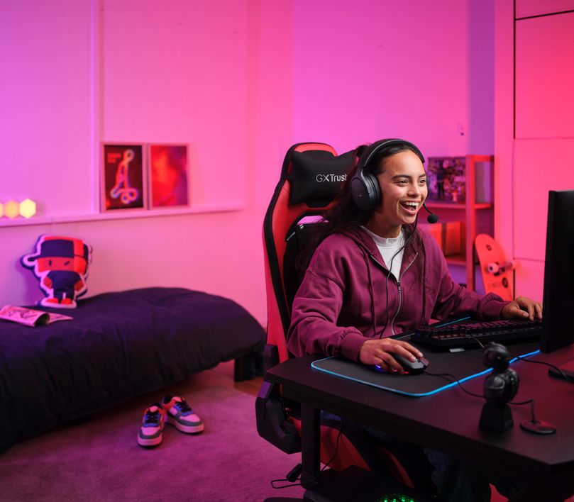 La imagen muestra a una persona sentada frente a una computadora con iluminación rosa. Lleva Auriculares Gaming Multiplataforma Trust GXT 489 Fayzo y está sentada en una silla de juego GXT Trust. La habitación tiene una cama al fondo y está decorada con elementos de temática gamer.