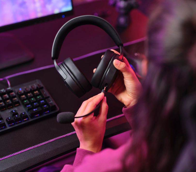 Una persona conectando el micrófono a unos Auriculares Gaming Multiplataforma Trust GXT 489 Fayzo cerca de un teclado y una computadora, con iluminación tenue y colores violetas en el fondo.
