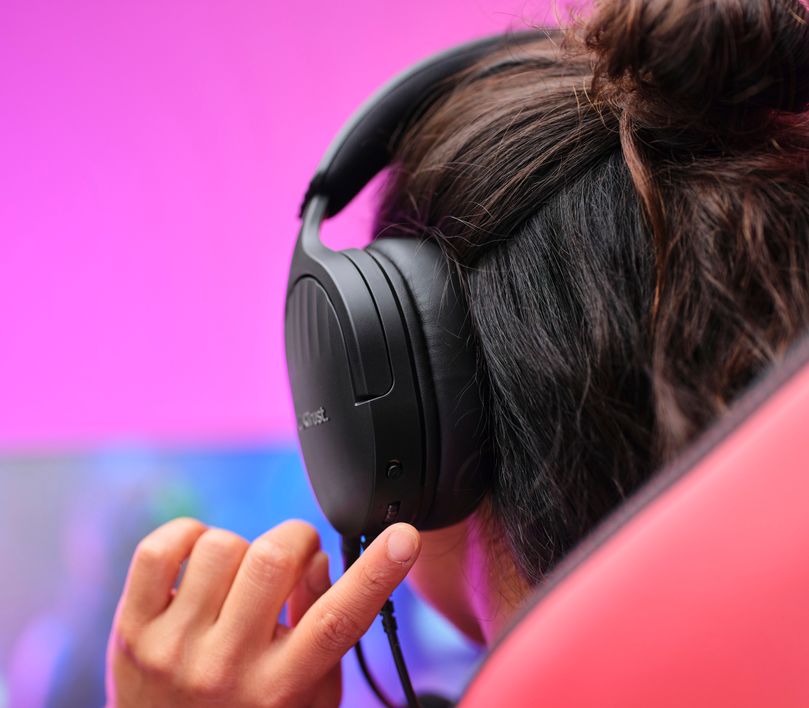 Una persona con Auriculares Gaming Multiplataforma Trust GXT 489 Fayzo negros está ajustando el volumen en un entorno con fondo rosa y azul.