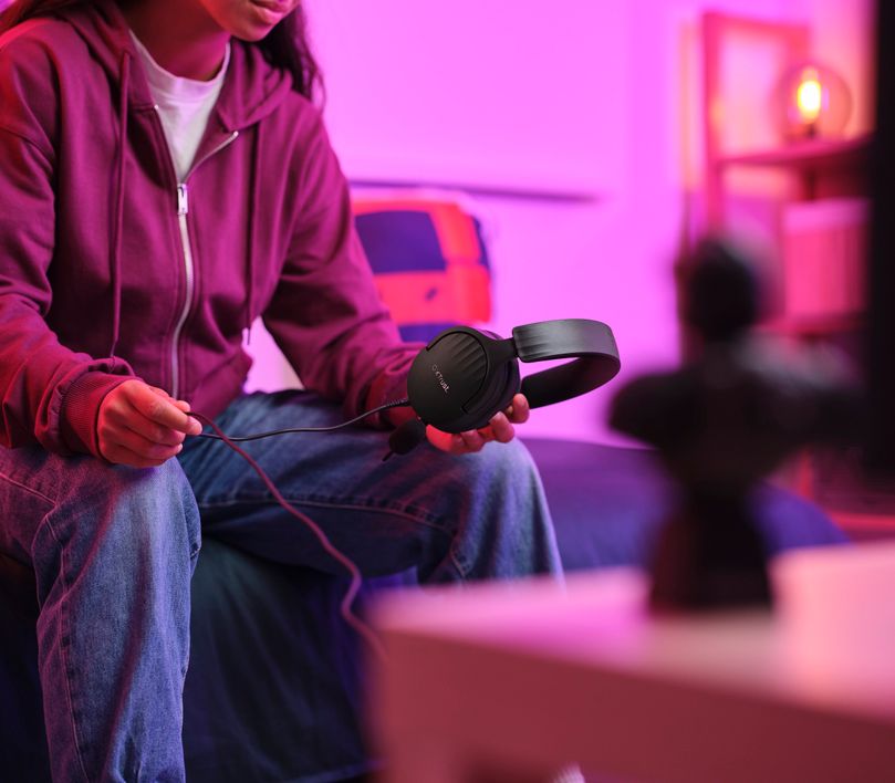 Una persona sostiene unos Auriculares Gaming Multiplataforma Trust GXT 489 Fayzo en una habitación iluminada con luces rosadas y violetas, con una figura borrosa en primer plano.