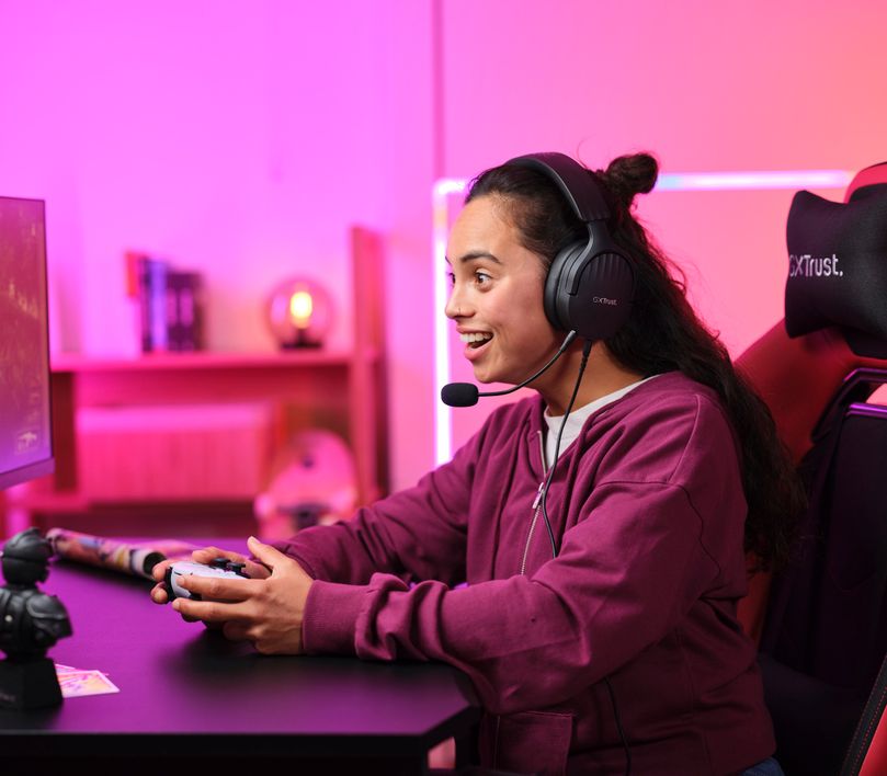 Una persona está jugando videojuegos con un controlador en una habitación iluminada con luces rosadas. Está sentada frente a un monitor y utiliza unos Auriculares Gaming Multiplataforma Trust GXT 489 Fayzo.