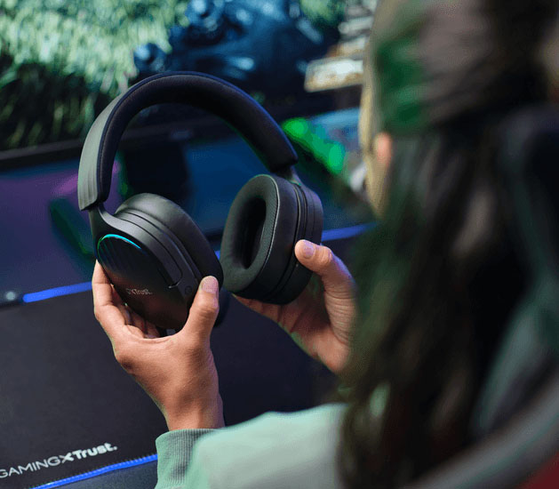 Manos sosteniendo Auriculares Gaming 7.1 USB Trust GXT 490 Fayzo frente a un monitor con un juego, sobre un escritorio con mousepad GAMINGxTRUST.