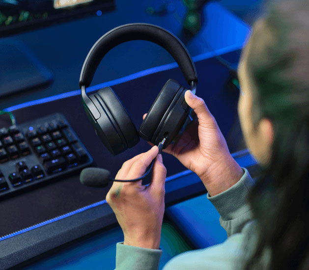 Una persona enchufando el micrófono a unos Auriculares Gaming 7.1 USB Trust GXT 490 Fayzo, con parte de un teclado y mousepad en un escritorio con luces de colores.