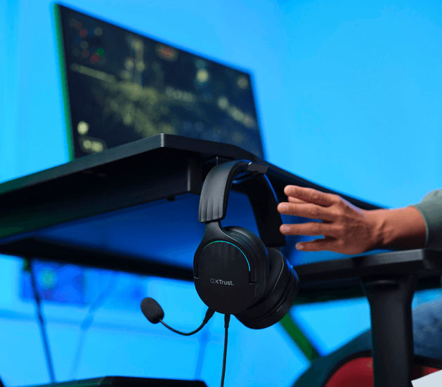 Auriculares Gaming 7.1 USB Trust GXT 490 Fayzo con micrófono colgando en un soporte sobre el borde de un escritorio, con una mano alcanzándolos y un monitor mostrando un juego de fondo.