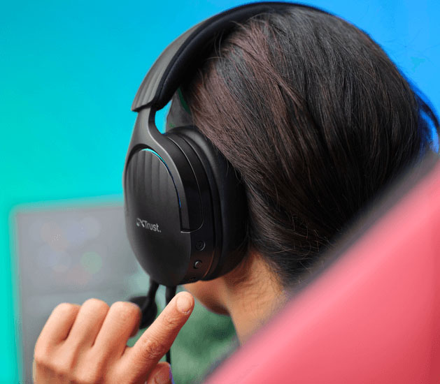 Primer plano de una persona usando Auriculares Gaming 7.1 USB Trust GXT 490 Fayzo negros, con una mano cerca, y un fondo de luces de colores desenfocado.