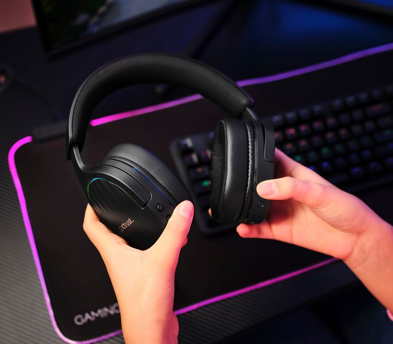 Una persona sosteniendo unos auriculares Gaming Inalámbricos Trust GXT 491 Fayzo, de color Negro sobre un escritorio con accesorios de gaming.
