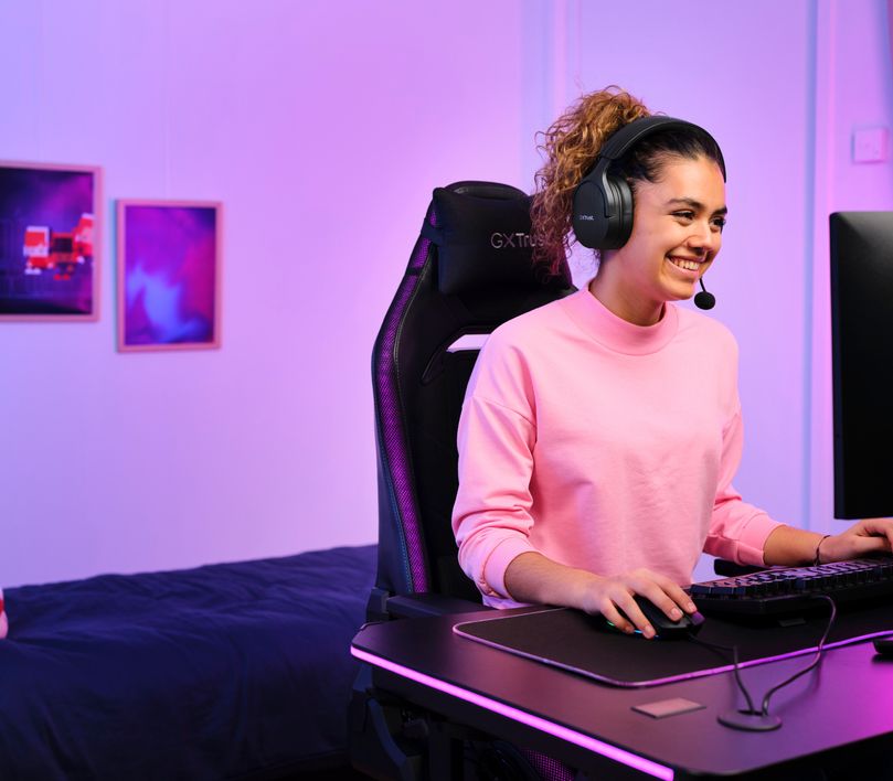 Persona sentada frente a una computadora con Auriculares Gaming Inalámbricos Trust GXT 491 Fayzo, de color Negro, jugando o trabajando en un ambiente iluminado con luces violetas.